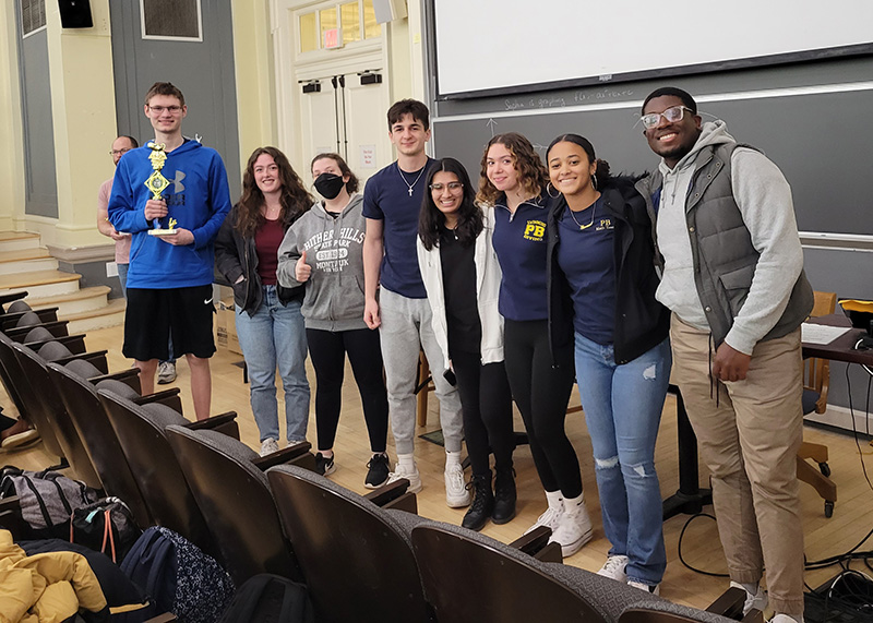 Eight high school students stand in a row on a stage smiling. the boy on the left is holding a trophy.