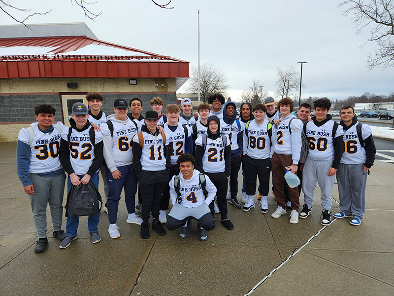 Twenty-one high school football players, all wearing white jerseys, stand together in front of a building.