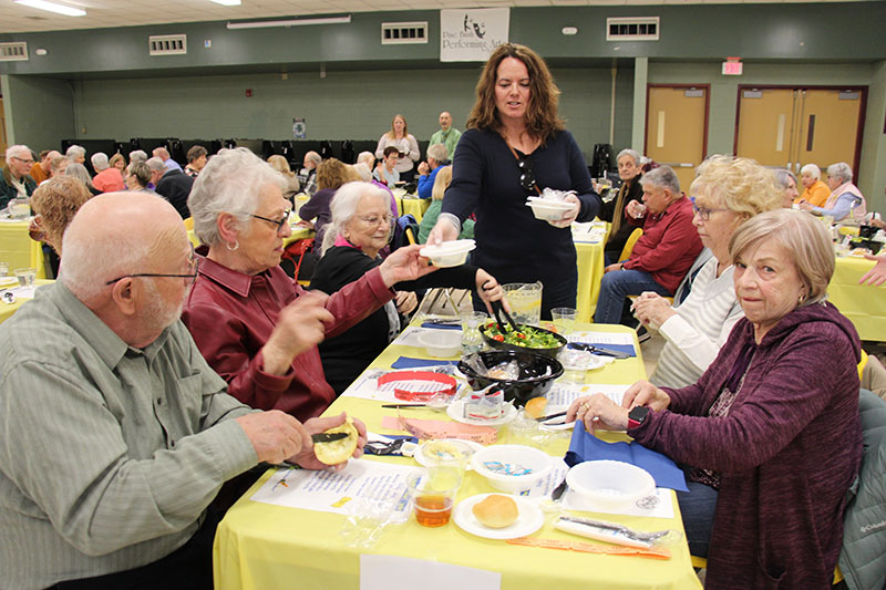 A group of six senior citizens sit at a table. There is a woman standing handing out dishes.