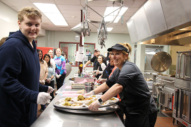 A group of people put food on plates and plates on large trays. All are smiling.