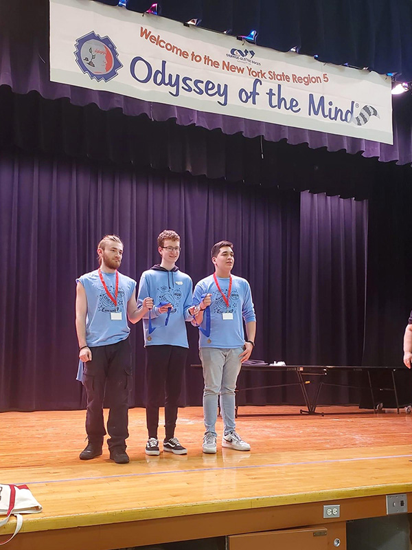 Three high school boys in light blue shirts stand on a stage with medals. Above them is a sign that says Odyssey of the Mind.