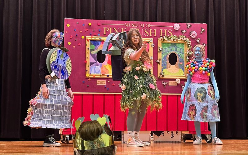 A group of three high school students dressed in costumes stand in front of a brightly colored fake wall they made for a project.