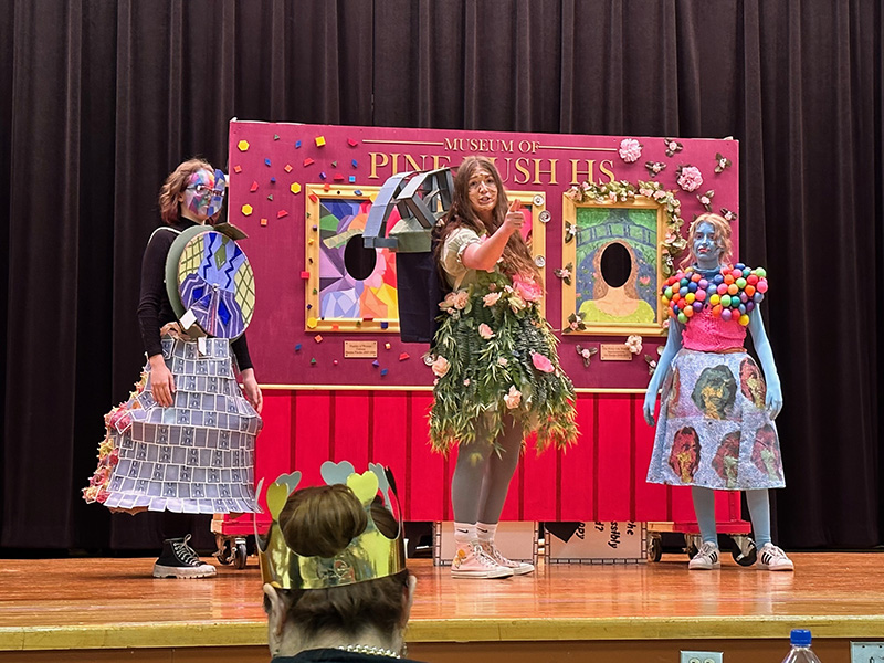 A group of three high school students dressed in costumes stand in front of a brightly colored fake wall they made for a project.
