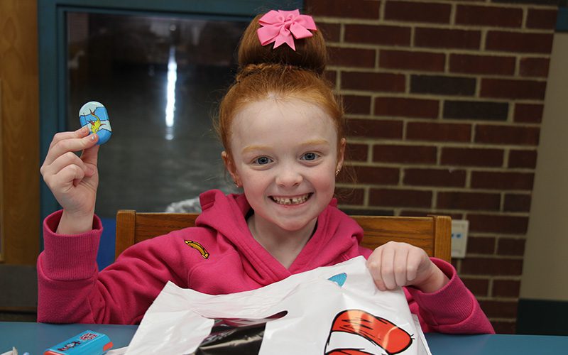 A kindergarten girl with red hair up in a large bun smiles holding her dr seuss bag and a little trinket with the dr. seuss fish on it.