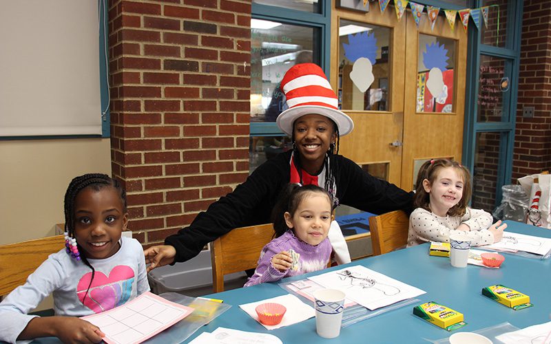 A high school girl dressed as the cat in the hat stands by three little kindergarten girls coloring and eating. They are all smiling.