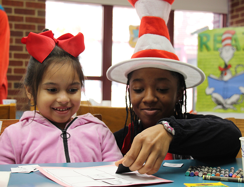A high school girl wearing a red and white striped hat plays tic tac toe with a kindergarten girl wearing a red bow and a pink jacket. Both are smiling.