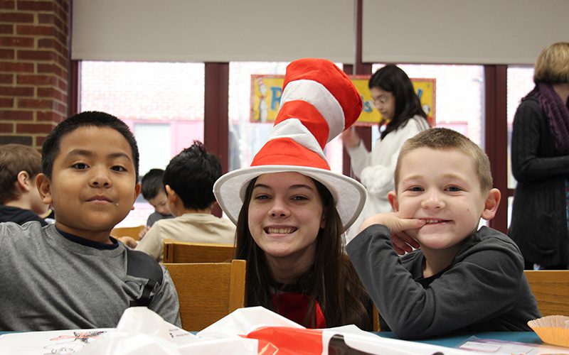 A high school girl dressed with a red and white striped tall hat sits with two kindergarten boys who are smiling.