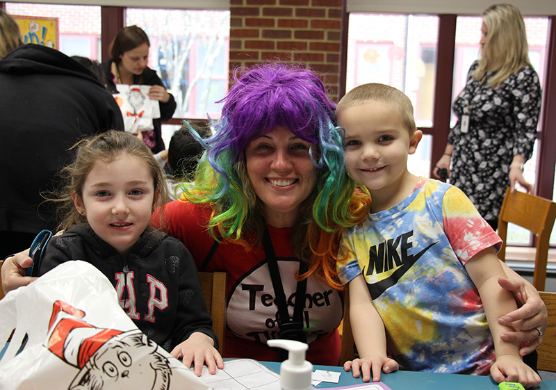 A woman wearing a red shirt with a large white circle on it and a rainbow colored wig smiles with two kindergarten students who are also smiling.