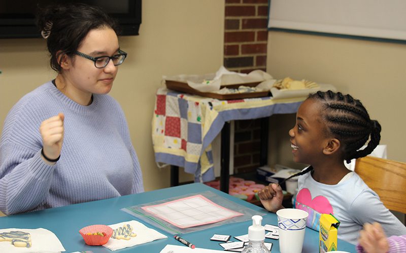 A high school girl with glasses and dark hair plays a game with a kindergarten student who is smiling broadly. She has dark hair in braids and wearing a light blue shirt.