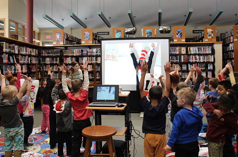 A high school student dressed as the cat in the hat stands in front of a room filled with kindergarten students with her hands up. the students have their hands up too. they are playing Simon Sez.