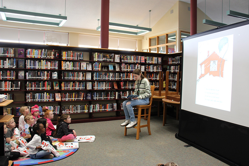 A group of kindergarten students sit on the floor listening as a high school student sitting on a chair reads a story to them. There is a screen behind her with the book she is reading projected on it.