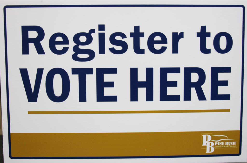 A white background with blue letters that says Register to VOTE HERE. There is a gold stripe on the bottom with the Pine Bush logo.