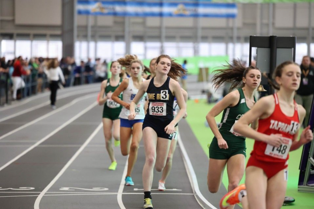 A high school young woman runs in a track meet.. She is wearing a navy blue shirt and shorts with Pine Bush written across the chest. There are other runners around her.