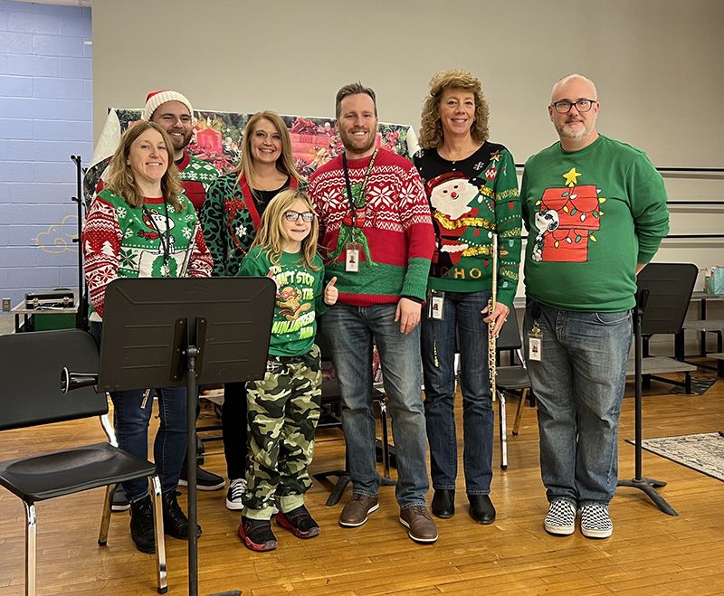 A group of adults - six of them - and one elementary student standing together. Some are holding musical instruments. They are on a stage.