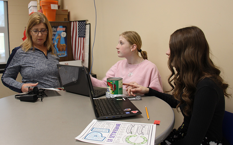 Two middle school girls sit at a table with their chromebooks. Their teacher stands with them looking at a piece of paper.