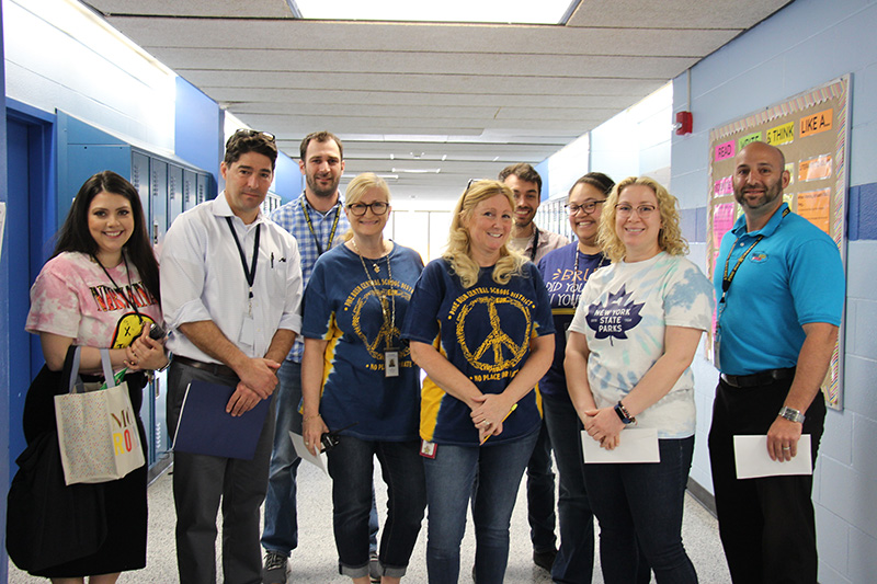 Nine adults stand together in a school hall smiling.