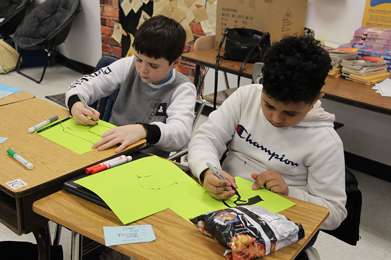 Two middle school boys sit at desks and create labels for a recycle bin.