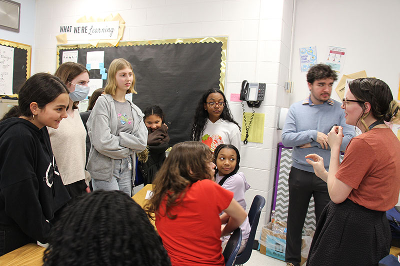 A group of middle school students stand in a group while a woman, their teacher, talks.