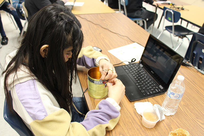 A girl with long dark hair uses a tape measure to measure the circumference of a can on a desk.