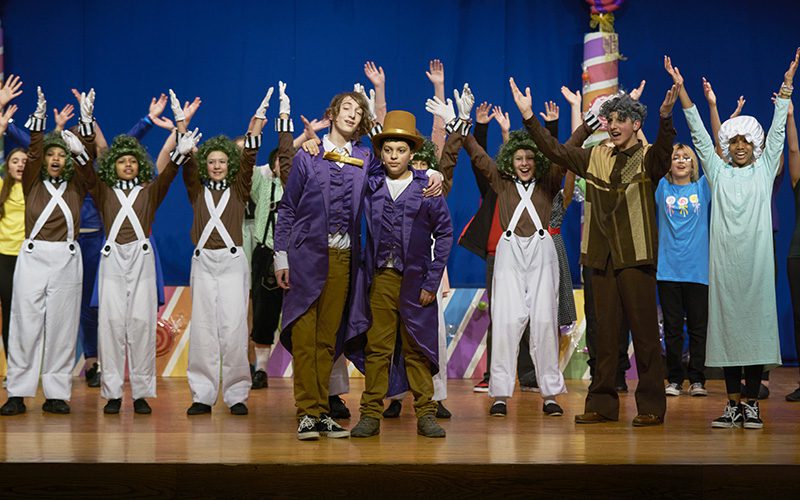 A group of about 1`2 middle school students dressed in costume for the Willy Wonka musical. They all have their arms up over their heads.