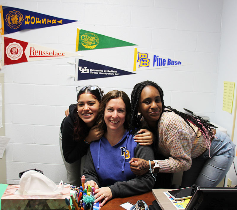 Two young women flank a woman who is sitting at her desk. There are collegiate pennants on the wall behind them. All are smiling.
