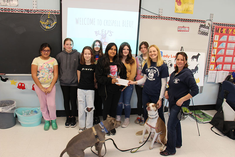 A group of six middle school dstudents stand with two adults and two dogs. One student is handing a check to one of the adults.