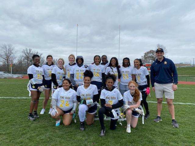 Fourteen high school girls dressed in shorts and white short-sleeve uniform shirts that say Pine bush in gold on them , stand with a man on the right. This is the flag football team.