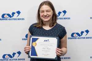 A young woman with shoulder-length dark blonde hair smiles. She is wearing a blue flowered short-sleeve shirt and holding a certificate. The background says Orange-Ulster BOCES.