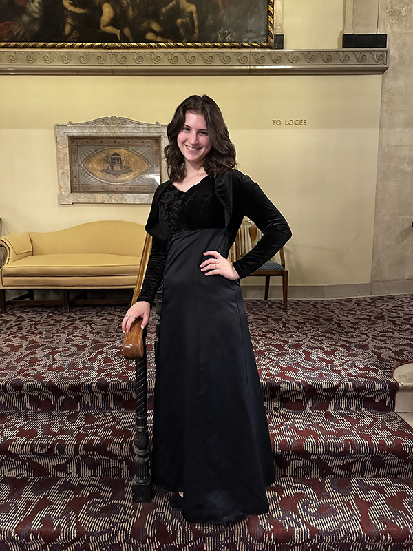 A young woman with shoulder-length dark hair wearing a long-sleeve black dress, stands on a step inside of a performance hall.