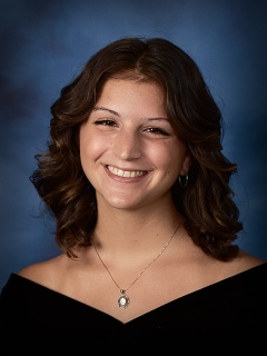 A traditional graduation photo of a young woman with shoulder-length dark hair smiling. She is wearing a v-neck, off the shoulder drape and a necklace.