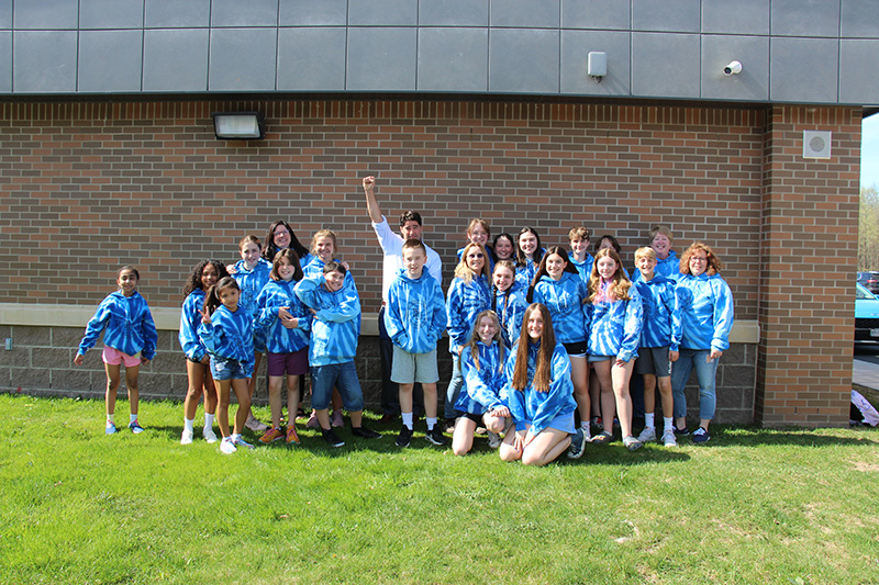 A large group of students - elementary, middle and high school - stand outside in front of a brick building, all wearing light and dark blue sweatshirts. There are four adults in the back with them. They are smiling, some are raising their arms in celebration.