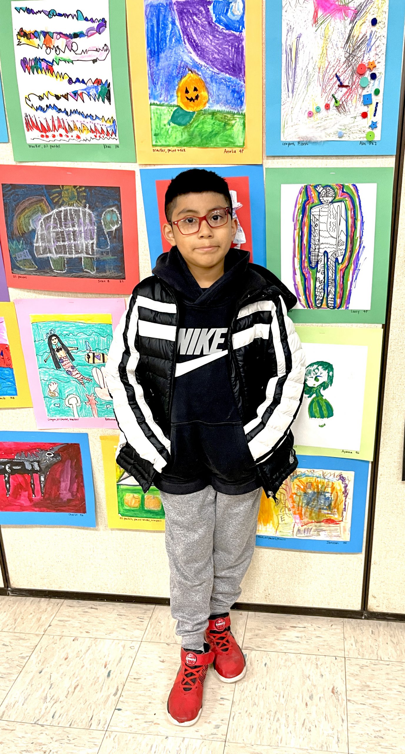 A boy wearing a black and white jacket and black shirt, glasses and dark hair  stands in front of a wall of artwork.