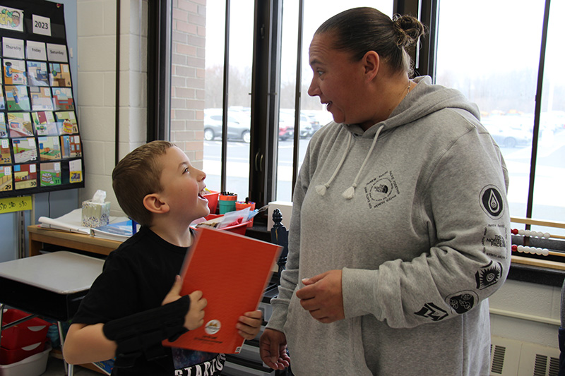 A second grade boy holds a red book in his hands and smiles looking up at an adult woman.