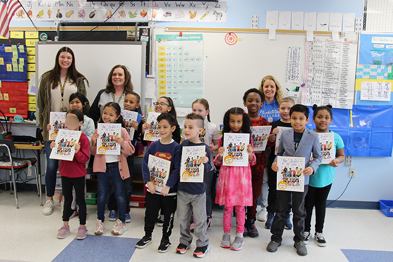 A class of 12 second grade students stand holding books they published. With them in the back are three women. All are smiling.