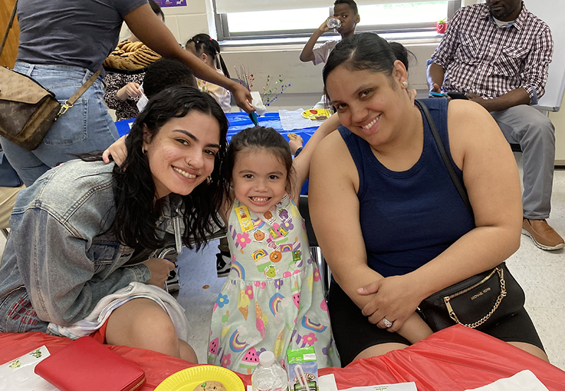 Two women smile as a little girl in a print dress stands between them, with arms around both of them. They are all smiling broadly.