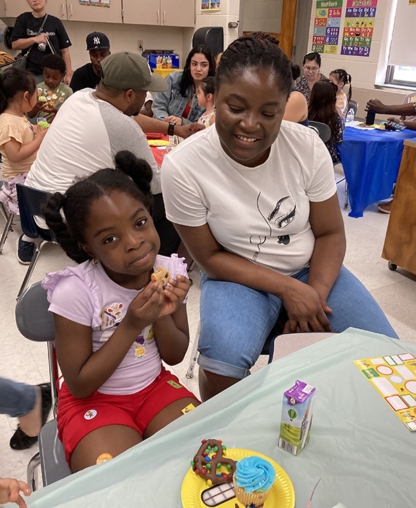 A little girl with her hair in pigtails eats a cookie as a woman sitting next to her looks on.