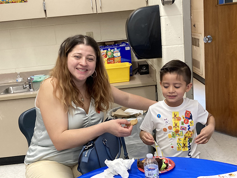 A kindergarten boy has his snack while a woman sitting next to him smiles and hands him food.