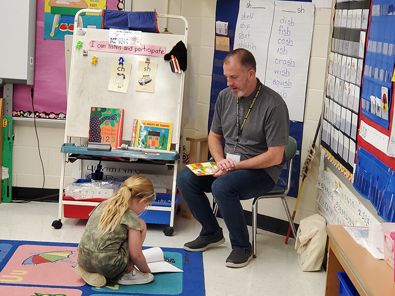 A kindergarten girl with long blonde hair sits on the floor reading her book to a man sitting in a chair.