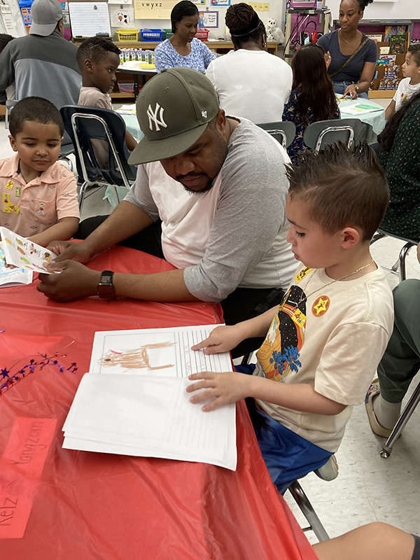 A boy sits at a table and reads his book he created to a man next to him.