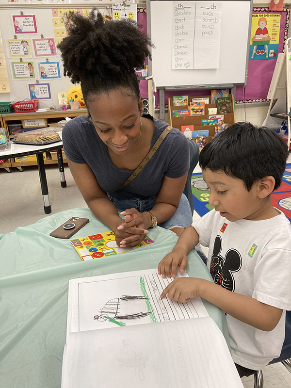 A kindergarten boy reads his book to a woman sitting next to him.