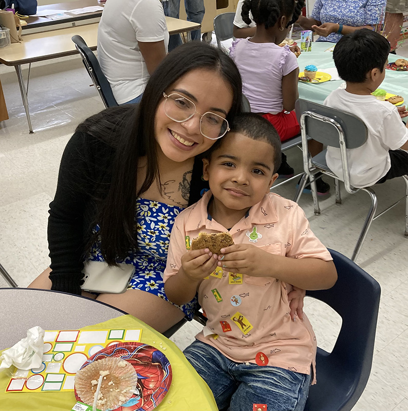 A woman and a kindergarten boy lean into each other for a picture. The woman has long dark hair and glasses and is wearing a blue shirt and black sweater. The boy has a peach colored shirt on and is holding a cookie in his hand.