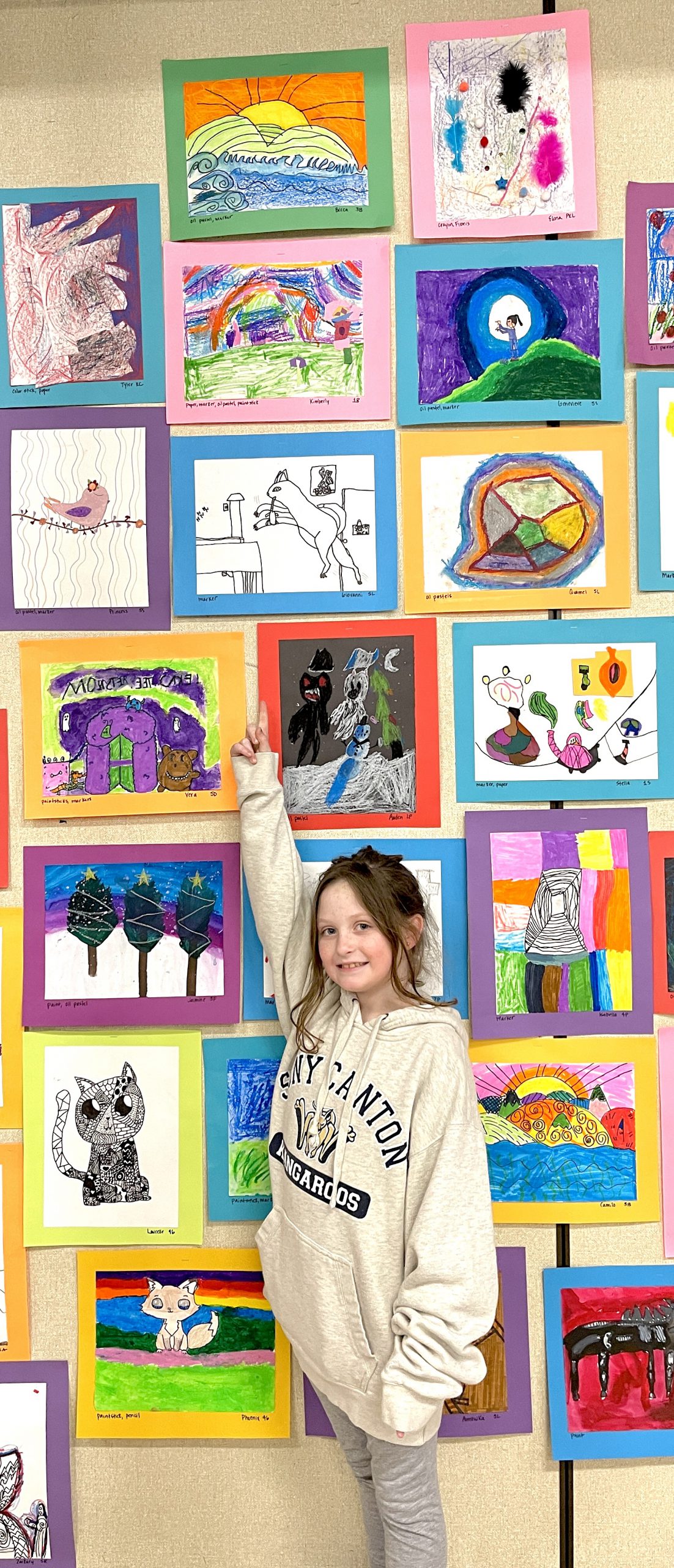 A girl with longer light hair, wearing a tan sweatshirt, points way up high to her piece of artwork high up on the wall.