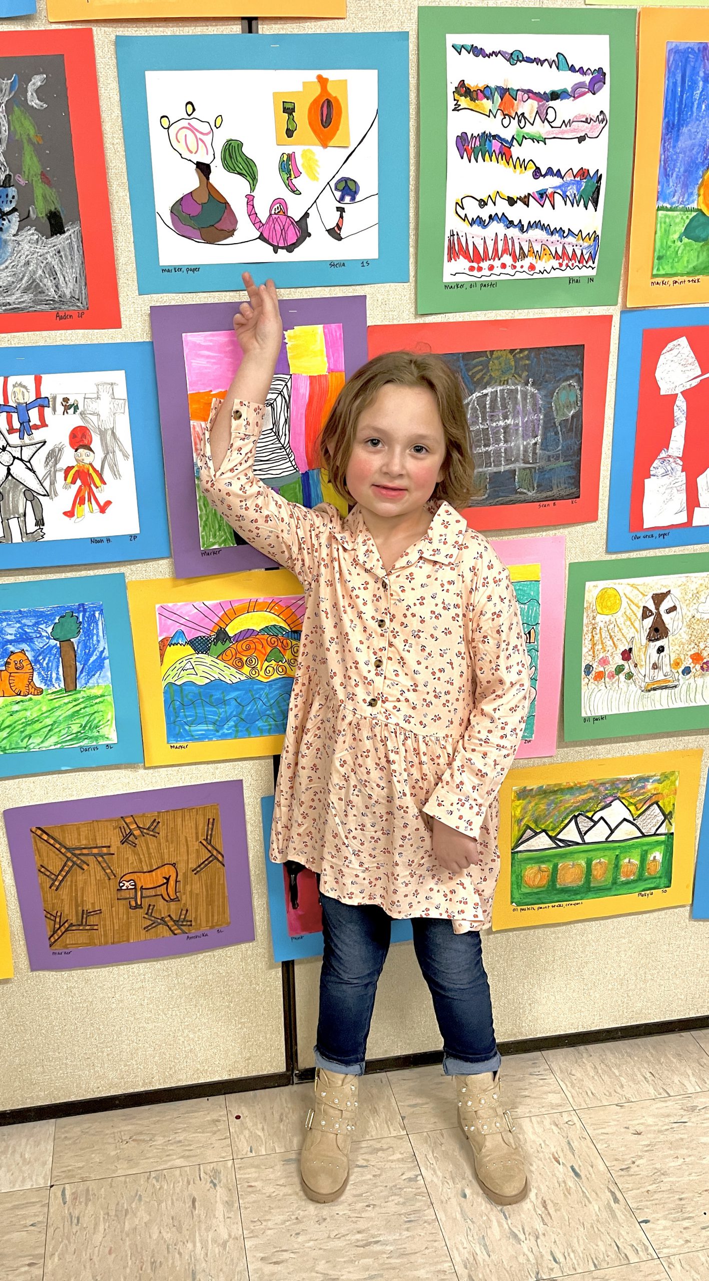A girl wearing jeans and a light colored printed  shirt stands in front of a wall of artwork points to her work.