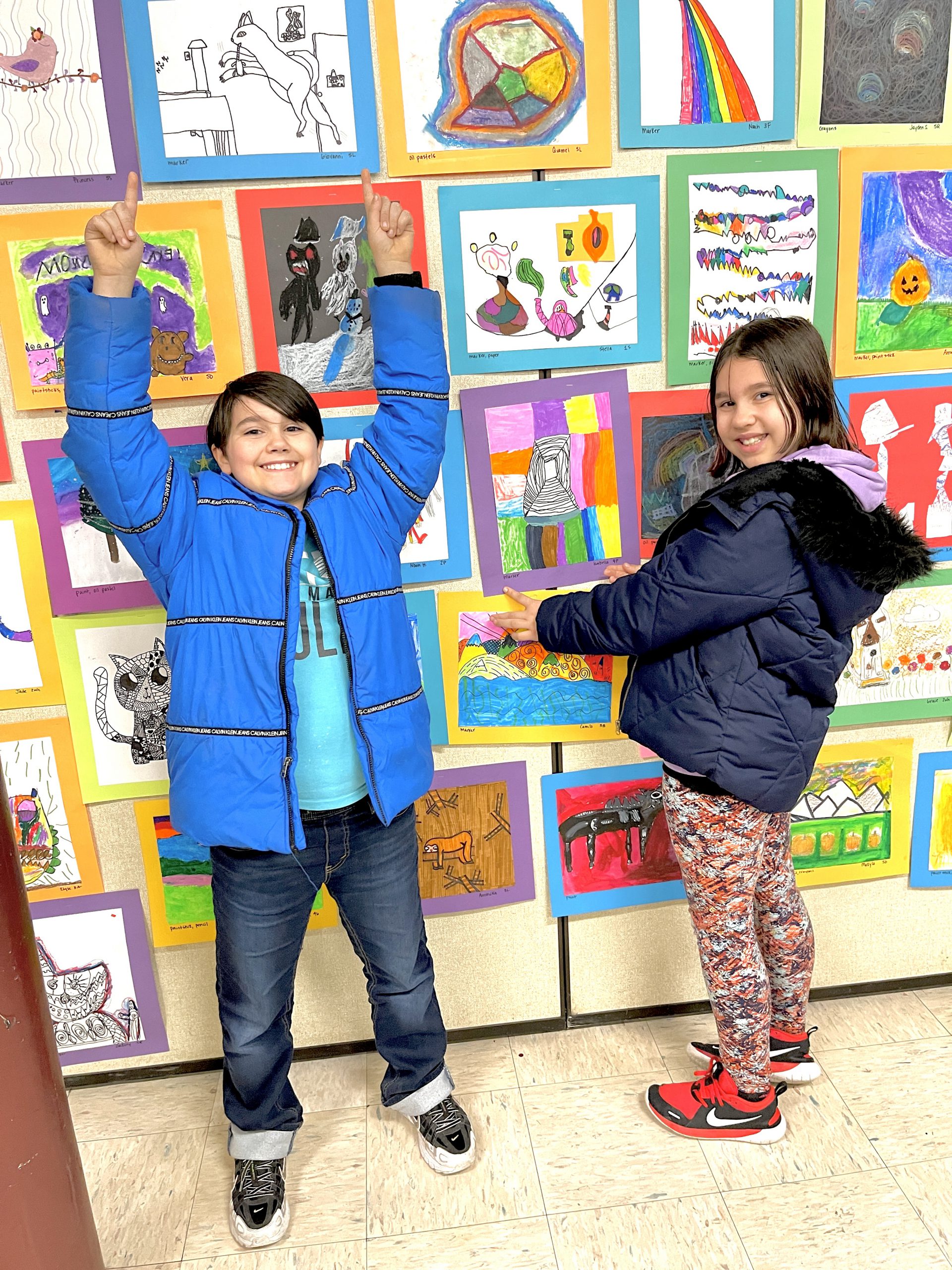 Two very happy elementary kids stand in front of a wall of artwork smiling and point to their art pieces.