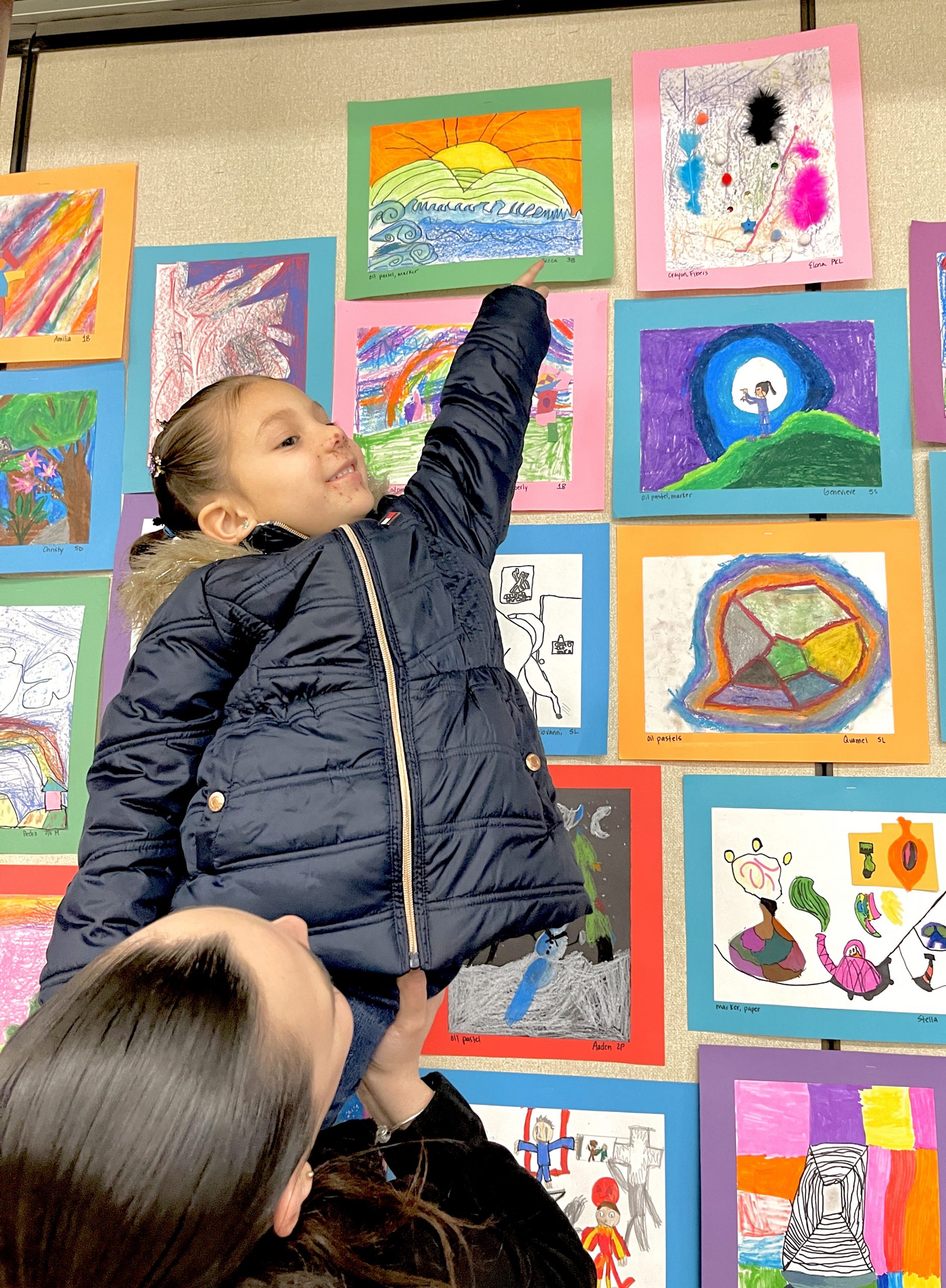 An elementary girl wearing a dark colored jacket points way up high on the wall as a woman holds her up. She is pointing to her artwork on the wall.
