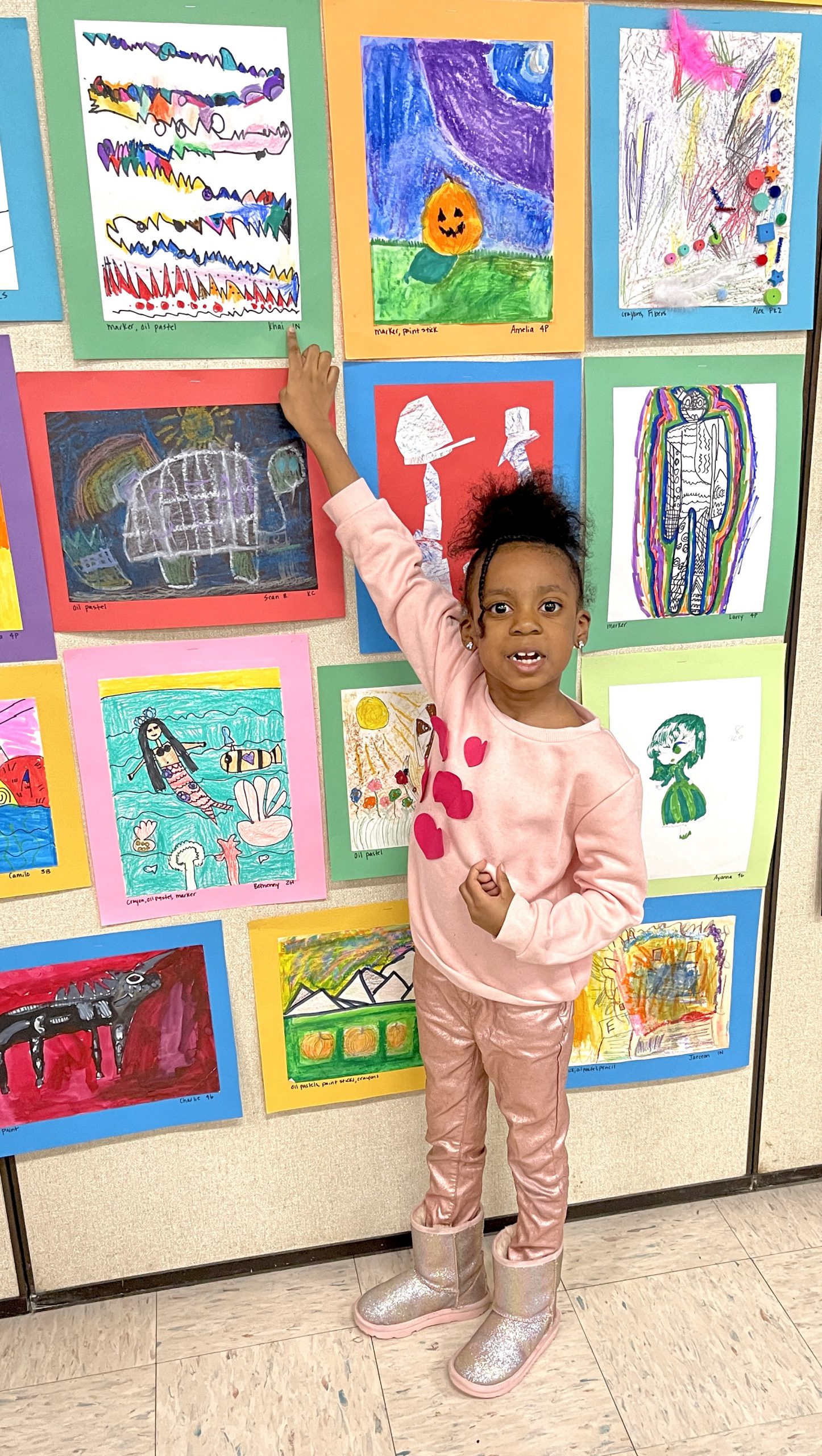 A little girl dressed in pink points to her artwork on the wall.