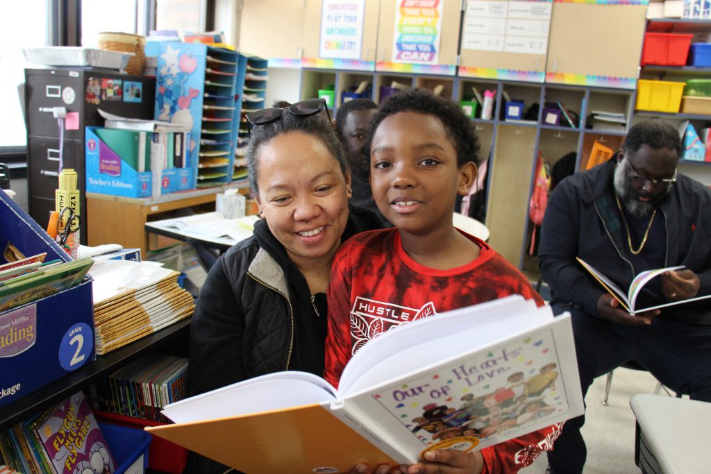 A second grade boy holds a book open and smiles as he reads with a woman.