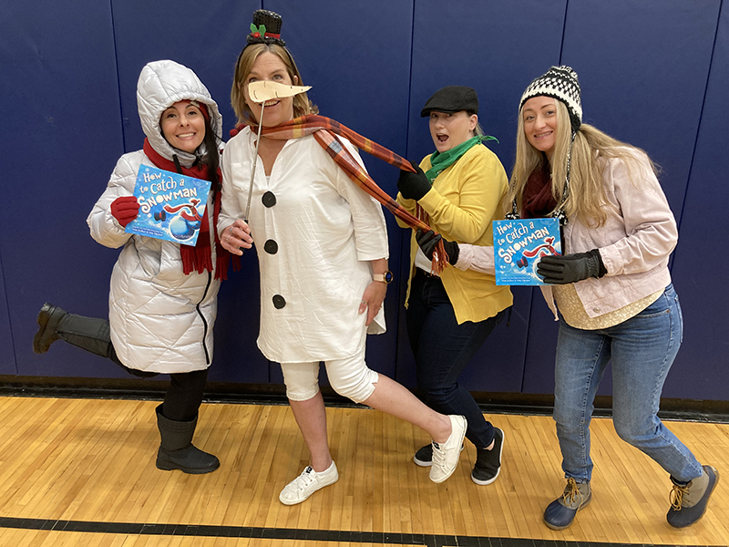 Four women dressed in winter clothing stand together holding a book How to Catch a Snowman 