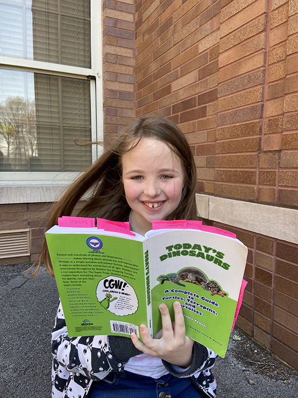 A girl smiles. She is outside reading a book called Today's dinosaurs. 