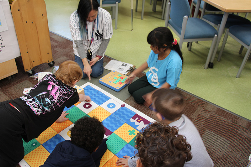 Five first grade students sit around a mat with patterns on it. There is a woman with long dark hair kneeling next to them.